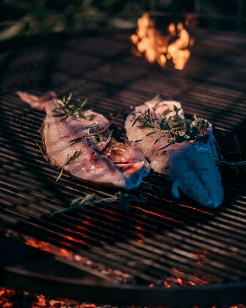 Zwei rohe Fischfilets mit Kräutern wie Rosmarin liegen auf einem Grillrost über glühender Holzkohle, im Hintergrund sind offene Flammen zu sehen.